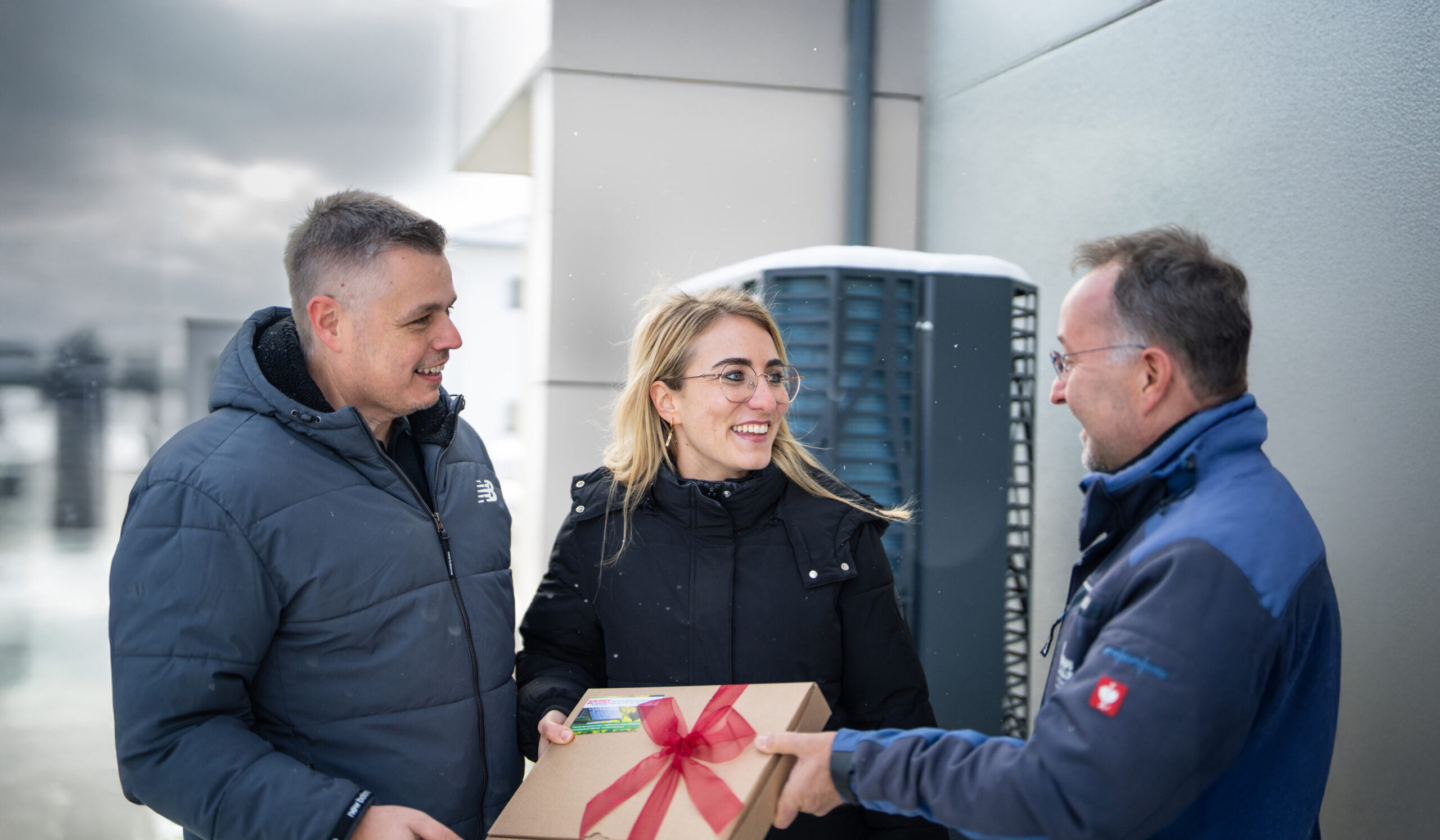 Drei lächelnde Personen bei der Übergabe eines Pakets mit Schleife